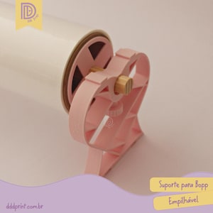 May include: A pink plastic Bopp film dispenser with a heart-shaped design. The dispenser holds a roll of white film and has a wooden dowel. The text "Suporte para Bopp Empilh&aacute;vel" is visible.