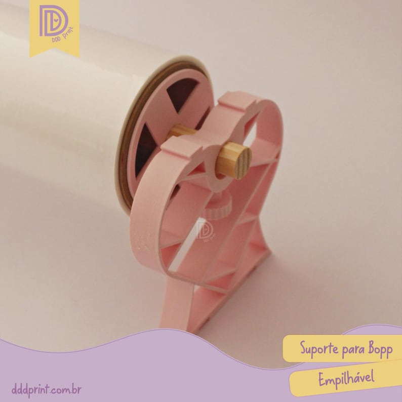 May include: A pink plastic Bopp film dispenser with a heart-shaped design. The dispenser holds a roll of white film and has a wooden dowel. The text "Suporte para Bopp Empilh&aacute;vel" is visible.