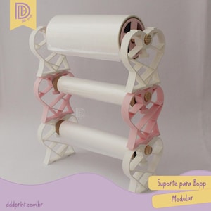 Pode incluir: Um suporte modular branco e rosa para rolos Bopp. O suporte tem um design em forma de coração e suporta três rolos de material branco. O texto "Suporte para Bopp Modular" é visível.