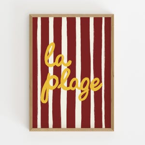 May include: A framed art print with a red and white striped background. The words "la plage" are written in yellow cursive script across the center of the image. The frame is a light brown color.