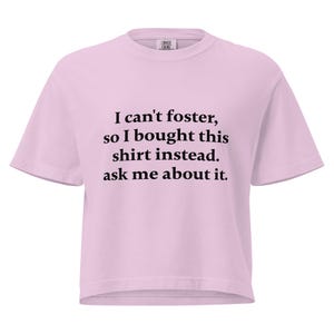 May include: A light pink t-shirt with short sleeves. The shirt features black text that reads, "I can't foster, so I bought this shirt instead. ask me about it."
