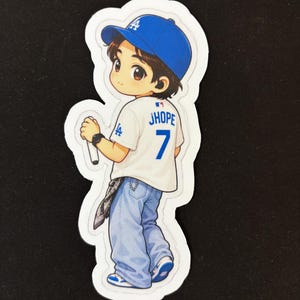 May include: A die-cut sticker featuring a cartoon character wearing a blue baseball cap with an LA logo, a white jersey with JHOPE and the number 7 on the back, and light blue jeans. The character holds a white microphone in one hand.