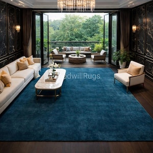 May include: A large, rectangular, teal blue rug dominates the room's interior. The rug is placed on a dark wood floor, and the room features white sofas, armchairs, and a marble coffee table. The space opens to a balcony with outdoor seating.