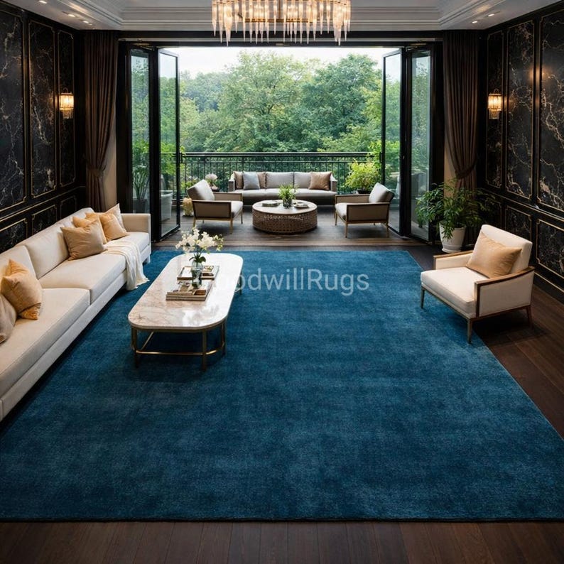 May include: A large, rectangular, teal blue rug dominates the room's interior. The rug is placed on a dark wood floor, and the room features white sofas, armchairs, and a marble coffee table. The space opens to a balcony with outdoor seating.