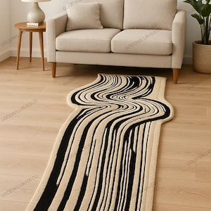 Può includere: Un tappeto astratto beige e nero con un design a strisce ondulate. Il tappeto è posizionato su un pavimento in legno davanti a un divano beige. Sono visibili anche un tavolino con una lampada e una pianta in vaso.