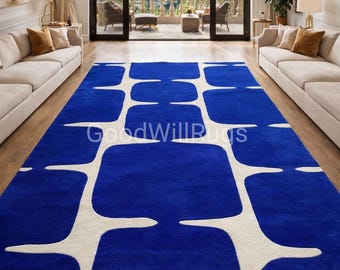 Puede incluir: Una alfombra azul rectangular larga con un patrón geométrico blanco. La alfombra está en una habitación espaciosa con grandes ventanales y vistas a palmeras. El texto "GoodWillRugs" es visible en la alfombra.