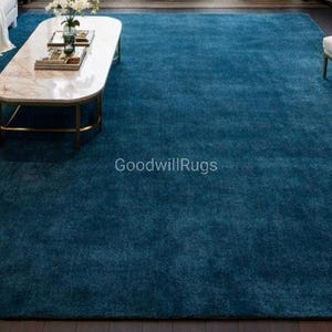 May include: A large, rectangular, teal-colored area rug dominates the room. The rug is placed under a white sofa, a marble-topped coffee table, and an armchair. The rug's texture appears soft and plush, adding warmth to the living space. The text "GoodwillRugs" is visible.