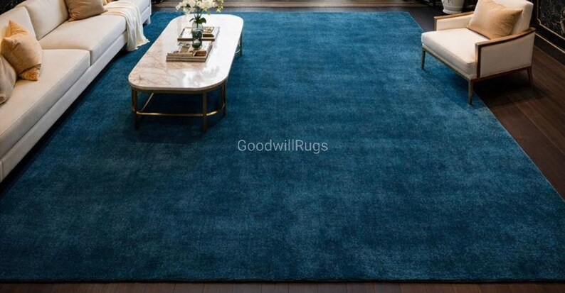 May include: A large, rectangular, teal-colored area rug dominates the room. The rug is placed under a white sofa, a marble-topped coffee table, and an armchair. The rug's texture appears soft and plush, adding warmth to the living space. The text "GoodwillRugs" is visible.
