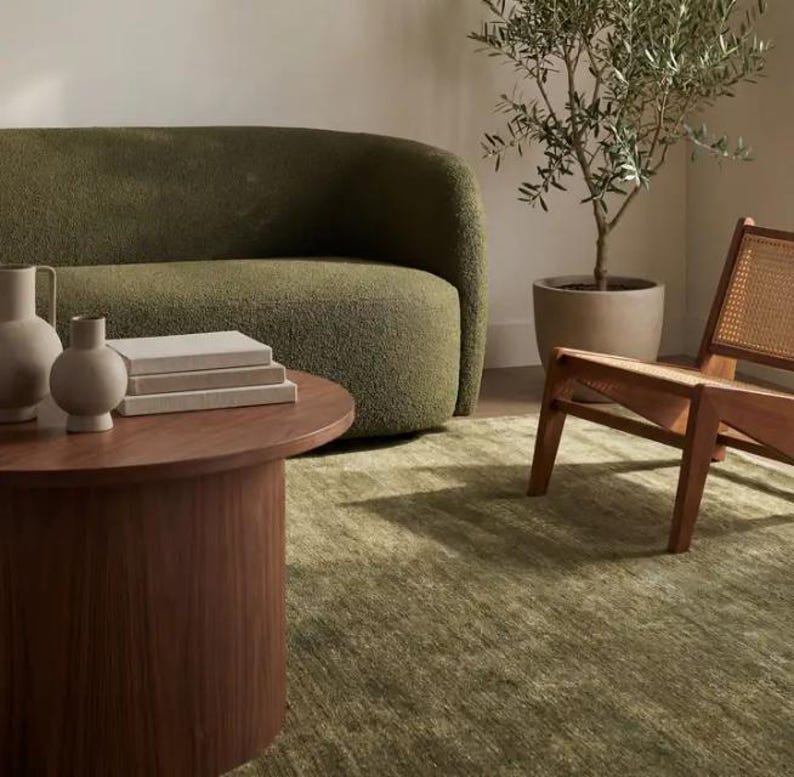 May include: A curved olive green textured sofa, a round wooden coffee table, two beige ceramic vases, a stack of books, a wooden chair with a woven cane back, and a potted olive tree in a beige planter, all arranged on a green area rug.