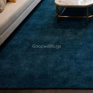 May include: A teal-colored area rug with a textured surface. The rug is placed in a living room setting, partially visible with a white sofa and a marble-topped coffee table. The text "GoodwillRugs" is printed on the rug.