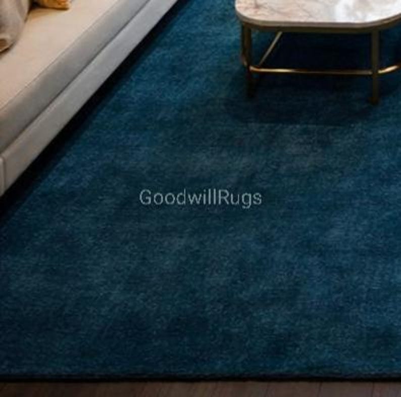 May include: A teal-colored area rug with a textured surface. The rug is placed in a living room setting, partially visible with a white sofa and a marble-topped coffee table. The text "GoodwillRugs" is printed on the rug.