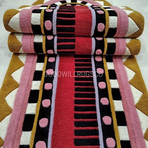 May include: Three rolled rugs with a geometric design. The rugs feature a color palette of red, pink, black, white, and gold. The text "GOODWILLRUGSCO" is visible on the bottom rug. The rugs are stacked on top of each other.