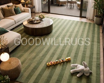 May include: A green striped rug with the text "GOODWILLRUGS" is the focal point of a living room. A toy train and a stuffed bunny are on the rug. The room has a white sofa, a round coffee table, and large windows.