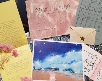 Puede incluir: Un conjunto de artículos de papelería que incluye una carta amarilla con flores rosas secas, un calendario Smile Tracker rosa, una tarjeta de Sudoku, un sobre pequeño con una pegatina de estrella, una lámina cuadrada y un dibujo de un conejo.