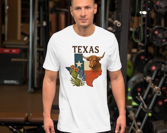 Op de afbeelding: Wit T-shirt met een Texas-afbeelding, inclusief een longhorn, cactus en blauwe bloemen. Het woord "TEXAS" staat in bruin boven de afbeelding. Het shirt heeft korte mouwen.