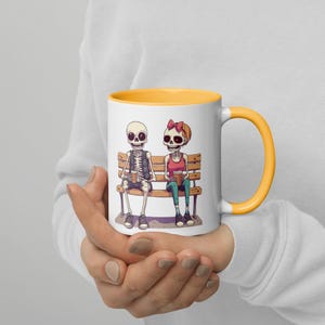 Puede incluir: Taza de cerámica blanca con interior y asa amarillos. La taza presenta una ilustración de dibujos animados de dos esqueletos sentados en un banco, sosteniendo tazas. Los esqueletos visten ropa y la esqueleto femenina tiene un lazo en el pelo.