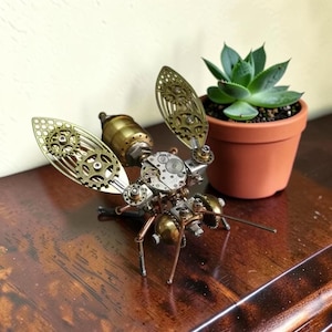 Op de afbeelding: Steampunk metalen insectenbeeld met messing en koperen onderdelen. De vleugels zijn gemaakt van ingewikkelde tandwielontwerpen. Het insect staat op een houten oppervlak, naast een vetplant in een pot.