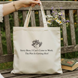 Könnte beinhalten: Cremefarbene Tragetasche mit dem Text "Sorry Boss, I Can't Come to Work. The Plot Is Getting Hot!" und einem floralen und Buch-Design. Die Tasche ist mit Blumen gefüllt.