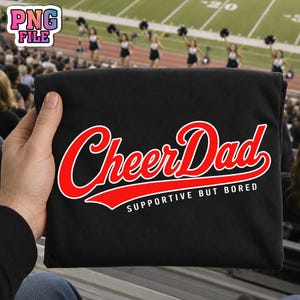 Op de afbeelding: Zwart T-shirt met de tekst "Cheer Dad" in rood script, omlijnd met wit. Daaronder staat de zin "Supportive But Bored" in het wit. Het shirt wordt omhoog gehouden tegen een stadionachtergrond.