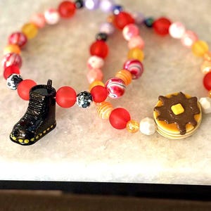 May include: Three beaded bracelets with colorful beads in shades of red, orange, yellow, and pink. One bracelet features a black boot charm, and another has a pancake charm with a pat of butter.