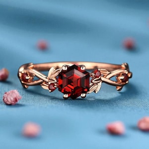 May include: Rose gold ring featuring a large, faceted, hexagonal red gemstone as the centerpiece. The ring is adorned with smaller red gemstones and delicate leaf and vine detailing. The ring is set against a soft blue background with scattered pink flower buds.