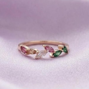May include: A gold ring with a leaf design, adorned with marquise-cut gemstones in pink, white, and green. The ring is set against a lavender background. A piece of jewellery for everyday wear.