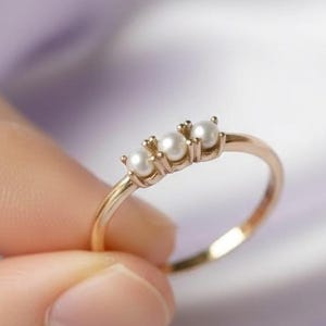Könnte beinhalten: Ein goldener Ring mit drei kleinen, runden weißen Perlen, die in einer horizontalen Reihe angeordnet sind. Das Ringband ist dünn und poliert, wobei die Perlen durch kleine Metallfassungen gehalten werden. Das Design ist schlicht.