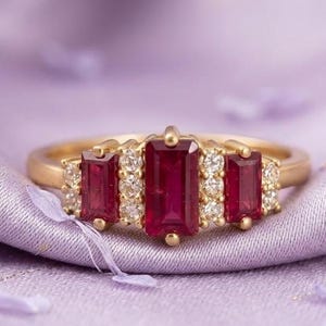 May include: A gold ring with three rectangular, deep red gemstones. The ring is accented with small, round, clear gemstones on either side of the larger stones. The ring is set against a soft, lavender fabric.