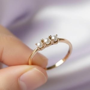 Op de afbeelding: Een dunne gouden ring met drie kleine, ronde, witte parels in een horizontale rij. De ringband is glad en gepolijst en toont een delicaat ontwerp waarbij de parels stevig op hun plaats worden gehouden door kleine metalen pootjes op een eenvoudige zetting.