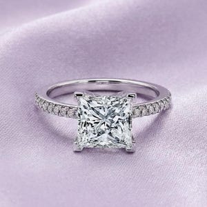 以下が含まれることがあります： 大きな正方形のダイヤモンドを中央に配した、シルバーの婚約指輪。バンドには小さなダイヤモンドがちりばめられています。指輪は柔らかいラベンダー色の布の上に置かれ、高級感を演出しています。