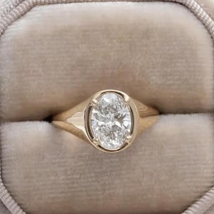 May include: A gold ring featuring an oval-cut diamond in a bezel setting, presented in a beige velvet ring box. The ring is centrally positioned, with a soft, blurred backdrop and delicate white flowers.