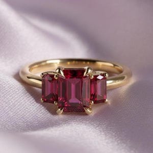 Puede incluir: Un anillo de oro con tres piedras preciosas rectangulares de color rojo intenso. La piedra central es más grande que las dos piedras laterales. El anillo está sobre una tela suave de color púrpura claro.