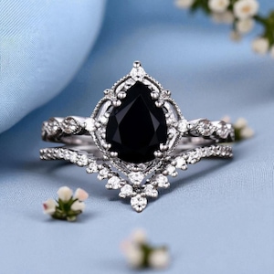 May include: A silver-toned ring set featuring a pear-shaped black gemstone. The engagement ring has a detailed filigree design around the center stone, and the band is adorned with small, clear accent stones. The wedding band has a curved design with small, clear accent stones.