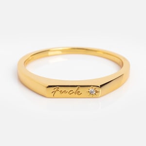 Puede incluir: Un anillo dorado con la palabra "fuck" grabada en la parte delantera. Una pequeña gema transparente está engastada junto a la palabra. El anillo tiene un diseño plano y rectangular.