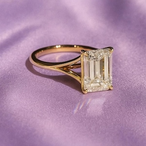 Könnte beinhalten: Ein goldener Verlobungsring mit einem großen, rechteckig geschliffenen Diamanten. Der Ring hat ein geteiltes Schaftband. Der Diamant ist klar und funkelnd, vor einem weichen, lila Hintergrund.