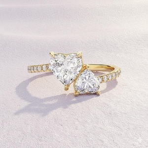 May include: A gold ring featuring two heart-shaped diamonds. The larger diamond is the focal point, with a smaller heart-shaped diamond adjacent. The band is adorned with smaller, round diamonds. The ring is set against a soft pink background.