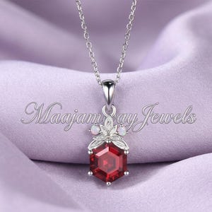May include: A silver necklace with a pendant featuring a large, faceted, red gemstone in a hexagonal shape. The pendant is adorned with smaller white gemstones and a floral design. The text "Maajam Jewels" is visible.