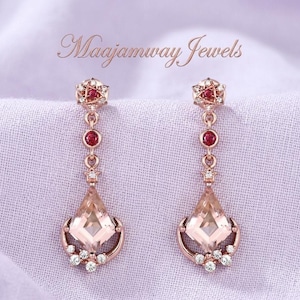 Op de afbeelding: Roségouden oorbellen met een geometrisch ontwerp. Elke oorbel heeft een stervormige bovenkant met rode accenten, een hangende druppelvormige steen en kleine witte edelstenen. De tekst "Maajamway Jewels" is zichtbaar.