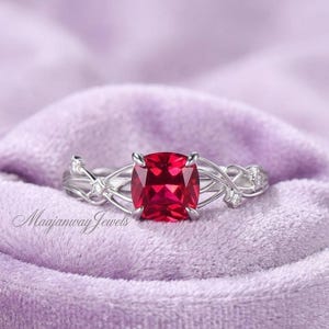 Puede incluir: Un anillo plateado con una gran gema roja brillante de talla cuadrada. La banda del anillo presenta un diseño delicado e entrelazado con pequeñas piedras de acento transparentes. El anillo está sobre un fondo morado suave.
