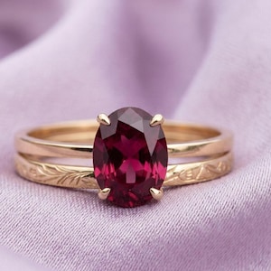 May include: A gold ring set with a large, oval-cut, deep red gemstone. The band is a simple gold band with a matching, engraved band. The ring is set against a soft, purple fabric.