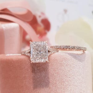 May include: A radiant-cut diamond engagement ring with a pave band, alongside a matching diamond wedding band. The rings rest on a pink velvet surface, with soft pink ribbons in the background. The rings sparkle brightly.
