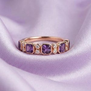 Puede incluir: Un anillo de oro rosa con piedras preciosas cuadradas moradas y pequeñas piedras preciosas redondas y transparentes. El anillo está sobre una tela lavanda suave. Las piedras están espaciadas uniformemente.
