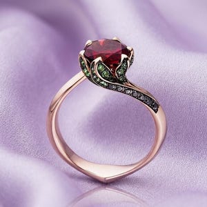 Könnte beinhalten: Ein roségoldener Ring mit einem großen, facettierten, tiefroten Edelstein. Die Fassung umfasst grüne und schwarze Akzentsteine mit einem fließenden, organischen Design. Der Ring wird auf einem weichen, lavendelfarbenen Stoff präsentiert.