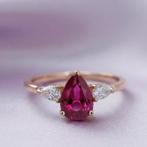 Peut inclure: Bague en or rose ornée d'une grosse pierre précieuse rose foncé en forme de poire. Deux petites pierres précieuses claires en forme de poire flanquent la pierre principale. Sur fond violet clair.