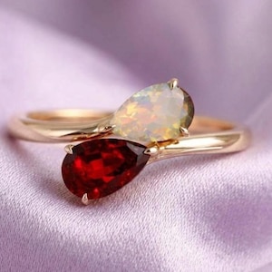 May include: A gold ring featuring two pear-shaped gemstones. One is a vibrant red, the other an iridescent opal. The ring's band curves gracefully, with the gemstones set opposite each other, creating a unique and elegant design. The ring is set against a soft, purple fabric.