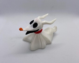 May include: A white figurine of Zero, the ghost dog from The Nightmare Before Christmas. The figurine has black eyes, a black mouth, and a small orange nose. It wears a red collar and has long, curved ears.