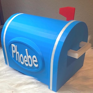 May include: A blue mailbox with white trim and a red flag. The name "Phoebe" is displayed in white lettering on the front of the mailbox. The mailbox has a handle and is sitting on a white surface.