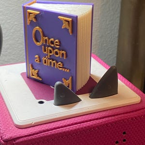 May include: A small purple book with gold detailing and the text "Once upon a time..." on the cover. It is supported by two grey triangular shapes on a white base.