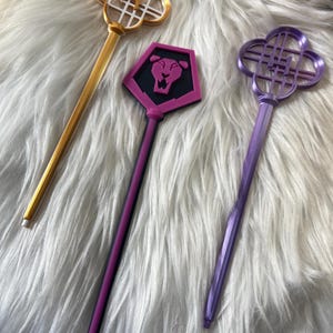 Op de afbeelding: Drie decoratieve toverstokken met unieke ontwerpen. Eén is goudkleurig met een klavervormige bovenkant, een andere is roze met een leeuwenembleem en de derde is paars met een klavervormige bovenkant. Allemaal hebben ze lange, slanke handvatten.