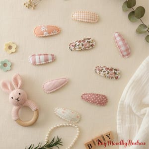 Pode incluir: Uma coleção de presilhas de cabelo para bebês em vários padrões e cores, incluindo floral, xadrez e bolinhas. Também há uma chocalho de coelho rosa de malha com um anel de madeira e um colar de pérolas. A estética geral é suave e em tons pastel.